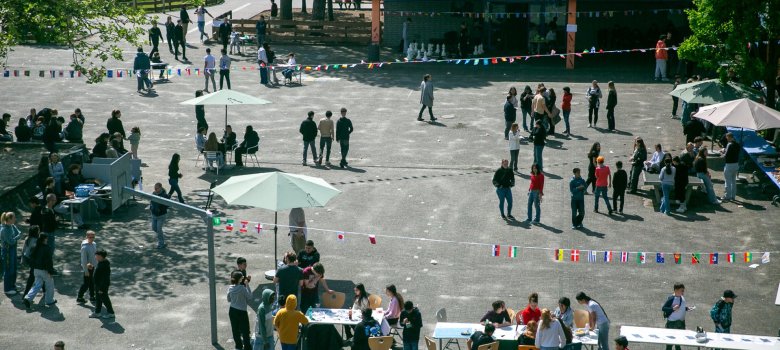 Das Foto zeigt den Schulhof aus erhöhter Perspektive. Über das Gelände verteilt stehen und gehen zahlreiche Schüler*innen zwischen Tischen, Ständen und Sitzgelegenheiten. Mehrere Sonnenschirme und Girlanden mit kleinen Länderflaggen sind zu sehen. Im Hintergrund befindet sich ein Schulgebäude.