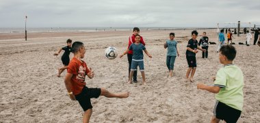 Mehrere Schülerinnen der Gesamtschule Gronau spielen auf dem breiten Sandstrand von Norderney Fußball. Ein Ball fliegt durch die Luft, während ein Schüler zum Schuss ansetzt und andere Mitspielerinnen sich auf dem Spielfeld verteilen. Im Hintergrund stehen ein Fußballtor, weitere Strandbesucher*innen sowie die Nordsee unter bewölktem Himmel.